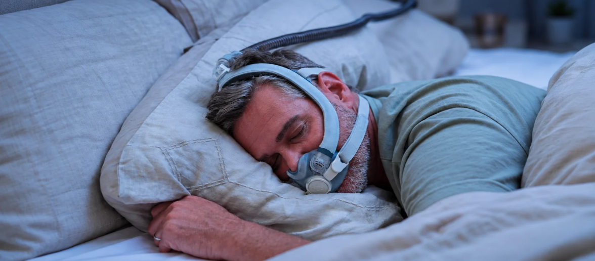 Middle-aged man sleeping on his side while wearing a CPAP mask connected to an air hose in a softly lit bedroom with cool blue nighttime lighting.